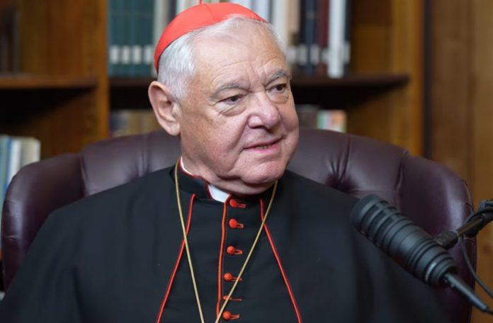 Cardinal in a black cassock with red trim and red skullcap, seated in a leather chair with a microphone nearby.”] ,