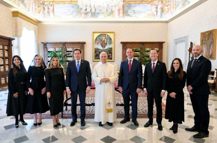 Eight adults in black formal attire stand with a man in white religious robes in a grand, chandelier-lit room with art and bookshelves.