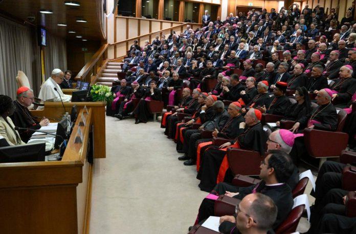 Pope at a podium speaking to a large hall filled with clergy in red and magenta attire and a seated audience behind him.