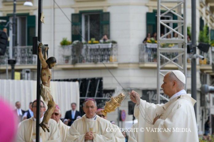 Catholic priest in white vestments blesses a suspended crucifix during an outdoor street procession, with a carved statue hanging from a pole nearby and onlookers in the background.