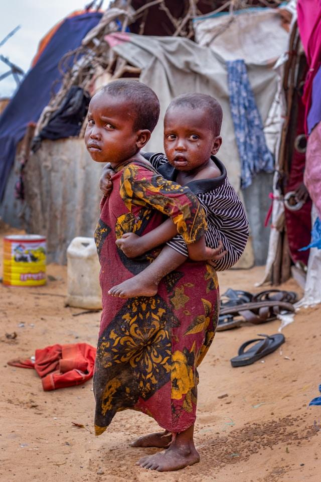 ismail-salad-osman-hajji-dirir-49K5LrGYbVo-unsplash Muno is 5 years old. She is carrying on her back her 2 year old baby brother. Two months ago, she and her family were displaced from the Bay region of Somalia after the drought killed all their livestock. Muno is not old enough to take responsibilities, but seems like the circumstances forced her to help her mother and take care of the younger child. (Photo by Ismail Salad Osman Hajji dirir on Unsplash)