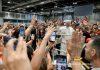 Pope Commends Volunteers in Madrid, Calling Selflessness the “Leaven” of a Better World Religious leader in white robes waves from a barrier to a cheering crowd holding up smartphones and tablets.
