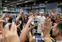 Pope Commends Volunteers in Madrid, Calling Selflessness the “Leaven” of a Better World Religious leader in white robes waves from a barrier to a cheering crowd holding up smartphones and tablets.