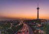 Iran’s Fragile Peace Deal Exposes Deep Divisions at Home and Abroad Evening cityscape with a busy highway and a tall illuminated tower on a hill at sunset.
