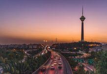 Iran’s Fragile Peace Deal Exposes Deep Divisions at Home and Abroad Evening cityscape with a busy highway and a tall illuminated tower on a hill at sunset.