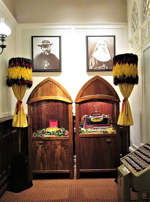 500px-Cathedral_Basilica_of_Our_Lady_of_Peace_interior_08 Two wooden arched display cases containing coffins and ceremonial items, flanked by tall feathered torches, with portraits on the wall behind.